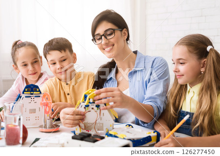 A teacher is assisting a group of students as they build robot models during a STEM classroom lesson. The students appear engaged and excited as they work on their projects 126277450