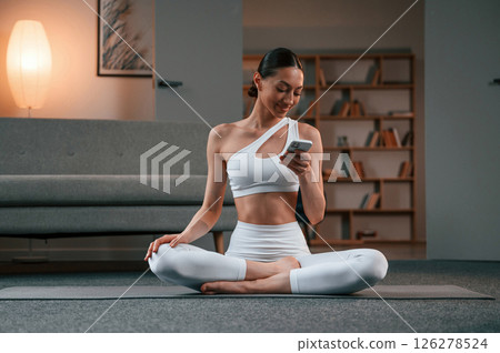 Holding smartphone. Young woman with slim body type in white sportswear is indoors doing yoga Holding smartphone. Young woman with slim body type in white sportswear is indoors doing yoga 126278524