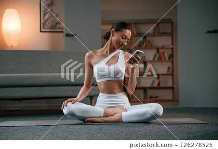 Holding smartphone. Young woman with slim body type in white sportswear is indoors doing yoga 126278525