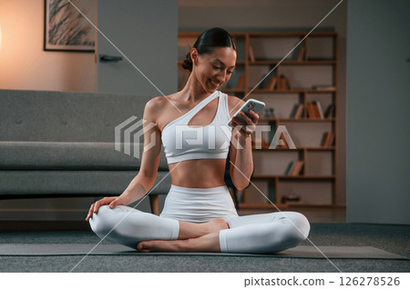 Holding smartphone. Young woman with slim body type in white sportswear is indoors doing yoga Holding smartphone. Young woman with slim body type in white sportswear is indoors doing yoga 126278526