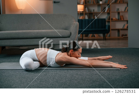 Stretching exercise. Young woman with slim body type in white sportswear is indoors doing yoga 126278531
