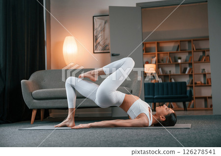 Exercise for legs and spine. Young woman with slim body type in white sportswear is indoors doing yoga Exercise for legs and spine. Young woman with slim body type in white sportswear is indoors doing yoga 126278541