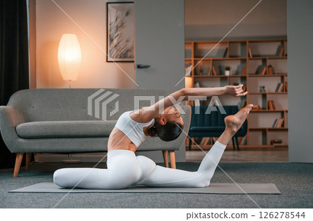 Practicing on the floor. Young woman with slim body type in white sportswear is indoors doing yoga 126278544