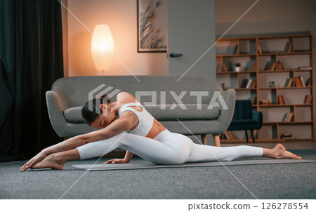 Exercise for legs and spine. Young woman with slim body type in white sportswear is indoors doing yoga Exercise for legs and spine. Young woman with slim body type in white sportswear is indoors doing yoga 126278554
