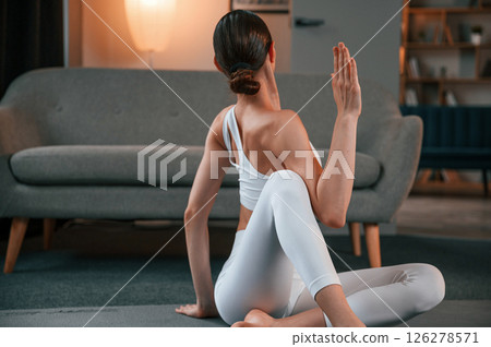Young woman with slim body type in white sportswear is indoors doing yoga 126278571