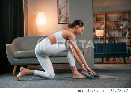 Lays out a yoga mat on the floor. Young woman with slim body type in white sportswear is indoors Lays out a yoga mat on the floor. Young woman with slim body type in white sportswear is indoors 126278572
