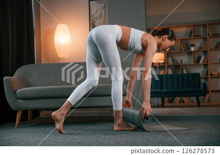 Lays out a yoga mat on the floor. Young woman with slim body type in white sportswear is indoors Lays out a yoga mat on the floor. Young woman with slim body type in white sportswear is indoors 126278573