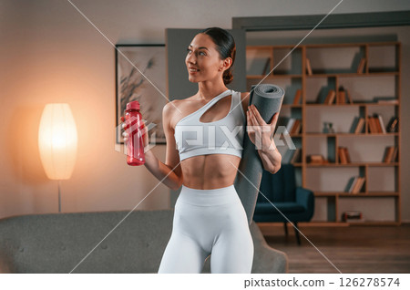With bottle of water. Young woman with slim body type in white sportswear is indoors with yoga mat 126278574
