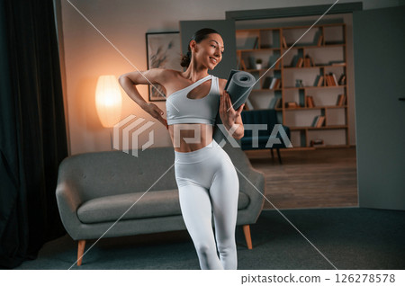 Standing and smiling. Young woman with slim body type in white sportswear is indoors with yoga mat 126278578