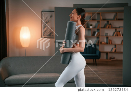 Woman with slim body type in white sportswear is indoors with yoga mat Woman with slim body type in white sportswear is indoors with yoga mat 126278584