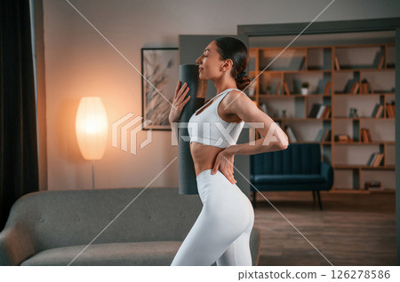 Ready for the session. Standing and holding yoga mat. Young woman with slim body type in white sportswear Ready for the session. Standing and holding yoga mat. Young woman with slim body type in white sportswear 126278586