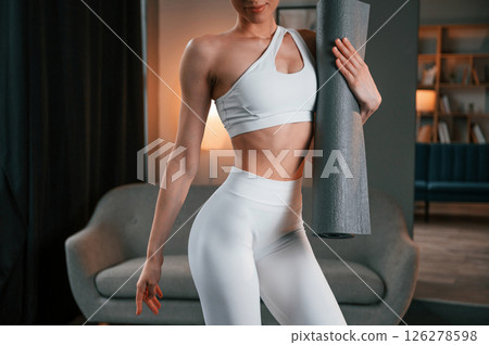 Close up view. Young woman with slim body type in white sportswear is indoors with yoga mat Close up view. Young woman with slim body type in white sportswear is indoors with yoga mat 126278598