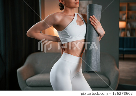 Showing the fitness type body. Young woman in white sportswear is indoors with yoga mat 126278602