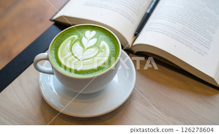 A cup of matcha latte with latte art sits beside an open book on a wooden table. A cup of matcha latte with latte art sits beside an open book on a wooden table. 126278604