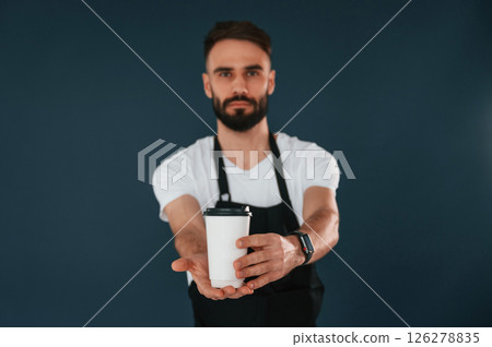 Giving the cup of coffee. Cafe worker is in apron. Handsome man is in the studio against blue background 126278835