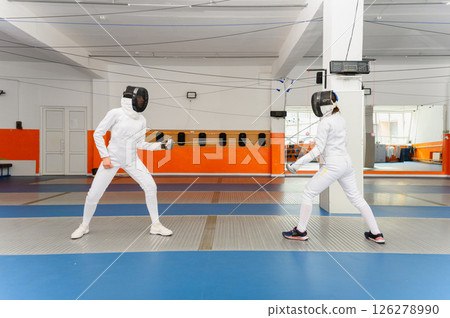 Two female fencers competing in bright indoor fencing hall. 126278990