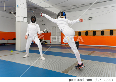Female fencer making airborne lunge attack in indoor duel. Female fencer making airborne lunge attack in indoor duel. 126279005