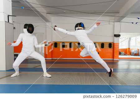 Female fencer making airborne lunge attack in indoor duel. Female fencer making airborne lunge attack in indoor duel. 126279009