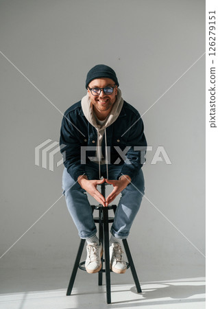 Model in hat and glasses is sitting on a chair. Handsome man is in the studio against white background 126279151