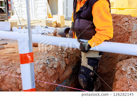 Construction worker fits PVC pipe into trench while surrounded by tools materials at site. Construction worker fits PVC pipe into trench while surrounded by tools materials at site. 126282145