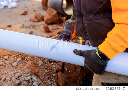 Skilled worker uses hand saw to cut plastic pipe at construction site, surrounded by materials. 126282147