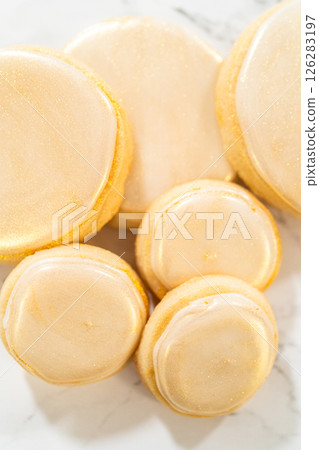 A collection of round sugar cookies decorated with smooth white royal icing and a shimmering gold dust finish. One cookie is being picked up, showing the delicate texture and elegant detailing. These 126283197