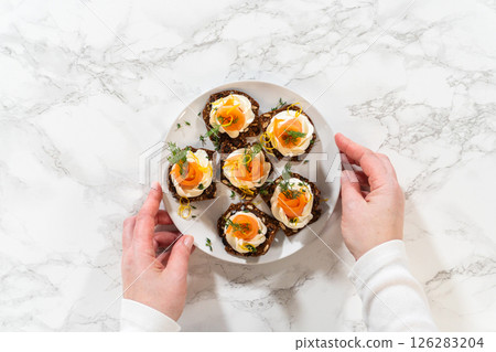 A top down view of a person holding a plate of smoked salmon canapes arranged on gourmet crispbread topped with cream cheese fresh herbs and lemon zest. The elegant appetizer is beautifully presented A top down view of a person holding a plate of smoked salmon canapes arranged on gourmet crispbread topped with cream cheese fresh herbs and lemon zest. The elegant appetizer is beautifully presented 126283204