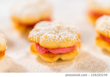 A stack of lemon spritz cookie sandwiches filled with cranberry buttercream, lightly dusted with powdered sugar and coconut flakes. The cookies have a delicate, buttery texture with a bright, festive 126283239