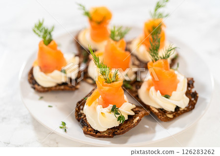A gourmet smoked salmon canape elegantly arranged on a crispy cracker, topped with a swirl of smooth cream cheese, fresh thyme, and a drizzle of olive oil. The vibrant orange salmon contrasts A gourmet smoked salmon canape elegantly arranged on a crispy cracker, topped with a swirl of smooth cream cheese, fresh thyme, and a drizzle of olive oil. The vibrant orange salmon contrasts 126283262
