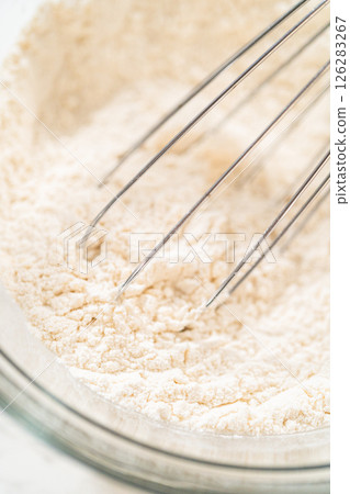 A glass mixing bowl filled with a blend of all-purpose flour, baking powder, salt, and cream of tartar, ready to be whisked together for making lemon spritz cookies. Combining dry ingredients 126283267