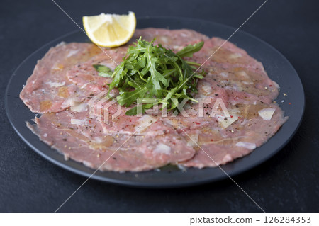 Marbled beef carpaccio with arugula, parmesan cheese, olive oil and lemon. A traditional classic appetizer made from thinly sliced raw meat. Close-up, selective focus, black background. 126284353