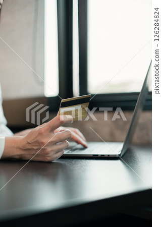 Close-up of a person holding a credit card while using a laptop for online shopping in a bright, modern workspace. 126284824