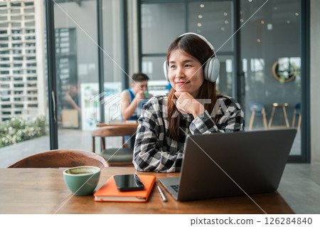 Young woman wearing headphones, working on a laptop in a modern cafe, smiling and enjoying a productive atmosphere. 126284840