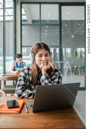 Smiling young woman wearing headphones, working on a laptop in a bright, modern cafe with large windows. 126284841