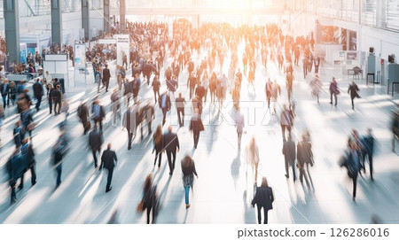 A blurry crowd of business people in a large, bright business hall. A dynamic scene unfolds as a blurry crowd of business professionals navigates the bright expanse of a large business hall. 126286016