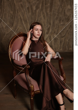 An Elegant Woman Dressed in a Beautiful Brown Dress Posing Gracefully on a Vintage Chair An Elegant Woman Dressed in a Beautiful Brown Dress Posing Gracefully on a Vintage Chair 126287433