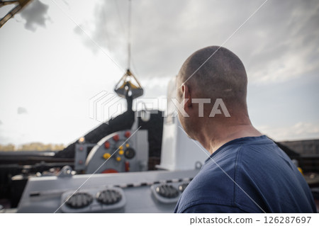 At the port, a worker oversees the process of loading coal onto a barge with the aid of a large crane. The setting features a sprawling river that flows gracefully alongside, reflecting the sun's rays 126287697