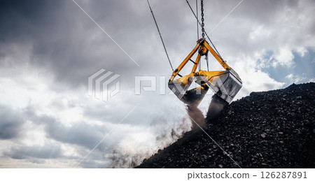 Excavator crane is engaged in loading coal using a large bucket, which is suspended and poised over a mound of black coal. Dust is billowing from the bucket as it lifts, indicating the heavy load 126287891