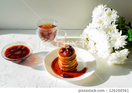 Fritters in a white plate with strawberry jam, a cup of tea and a bouquet of white chrysanthemums on a white table. Fritters in a white plate with strawberry jam, a cup of tea and a bouquet of white chrysanthemums on a white table. 126288561