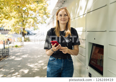 Young Woman Using Parcel Locker with Smartphone Outdoors 126289419