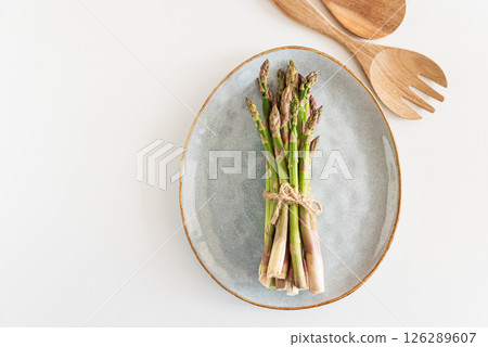 Fresh asparagus tied bundle on plate and wooden utensils on white table. Top view Fresh asparagus tied bundle on plate and wooden utensils on white table. Top view 126289607