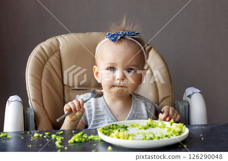 Cute baby girl with blue eyes eating messy food with spoon while sitting in high chair, learning to self-feed with broccoli puree, showing curiosity, innocence and healthy eating habits at home 126290048
