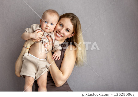 Portrait beautiful happy blonde mother with cute baby in beige clothing, smiling and looking at camera, isolated on neutral gray background, joyful family mom and child 126290062