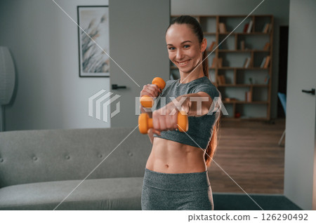 Positive young woman in yoga clothes is smiling and holding dumbbells 126290492