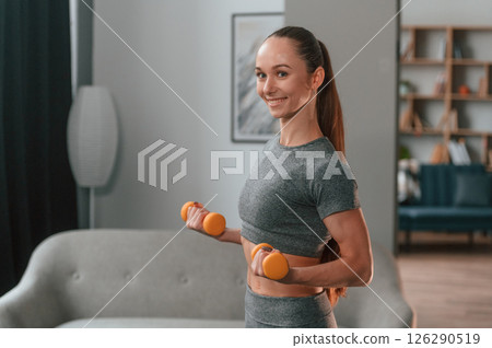 Yellow colored dumbbells in hands. Young woman in yoga clothes doing fitness indoors Yellow colored dumbbells in hands. Young woman in yoga clothes doing fitness indoors 126290519