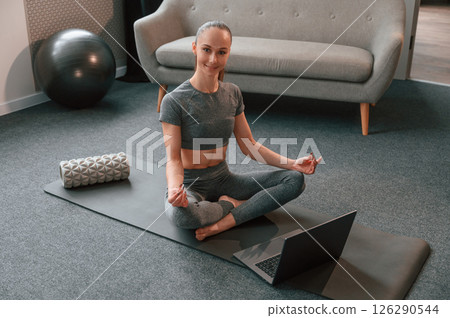 Taking online lessons by using laptop. Young woman in yoga clothes doing fitness indoors Taking online lessons by using laptop. Young woman in yoga clothes doing fitness indoors 126290544
