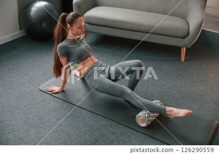 Legs exercises by using foam roller. Young woman in yoga clothes doing fitness indoors Legs exercises by using foam roller. Young woman in yoga clothes doing fitness indoors 126290559