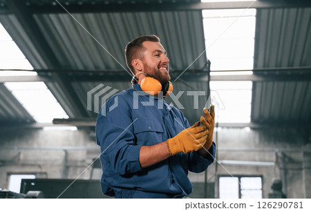 In yellow colored hear protective headphones and gloves. Factory worker in blue uniform is indoors In yellow colored hear protective headphones and gloves. Factory worker in blue uniform is indoors 126290781