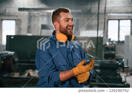 In yellow colored hear protective headphones and gloves. Factory worker in blue uniform is indoors 126290782