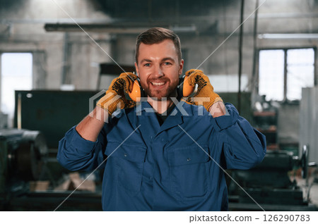 In yellow colored hear protective headphones and gloves. Factory worker in blue uniform is indoors 126290783